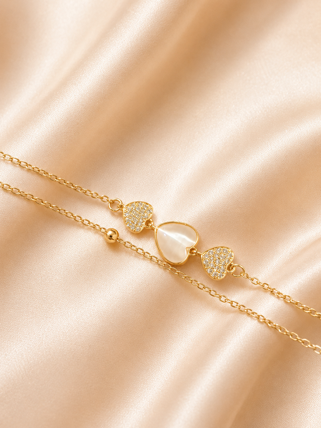 A delicate gold chain bracelet with three heart charms, featuring two pavé-set heart charms and one larger heart with a milky white inlay, displayed on a draped champagne satin fabric. The Whispering Hearts Bracelet by Ashaarya featuring 18KT gold plating, mother of pearl hearts, cubic zirconia, anti-tarnish 316L stainless steel comfort jewellery.