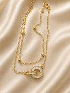 A gold-toned double-strand necklace with a circular pendant featuring clear stones, displayed on a beige satin fabric background. anti-tarnish, comfort jewellery, gold bracelet for women, layered bracelet for women, interlocking circle bracelet, lightweight bracelet, everyday bracelet, stone bracelet for women, meaningful jewellery, delicate gold bracelet, adjustable bracelet, gift for women, Ashaarya bracelet