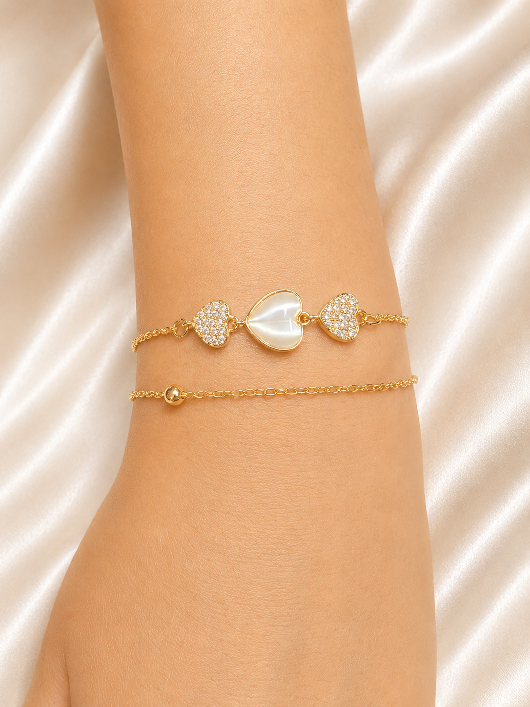 A close-up of a person's wrist wearing two delicate gold bracelets. The top bracelet features a central white mother-of-pearl heart flanked by two pavé-set heart charms. The Whispering Hearts Bracelet by Ashaarya featuring 18KT gold plating, mother of pearl hearts, cubic zirconia, anti-tarnish 316L stainless steel comfort jewellery.