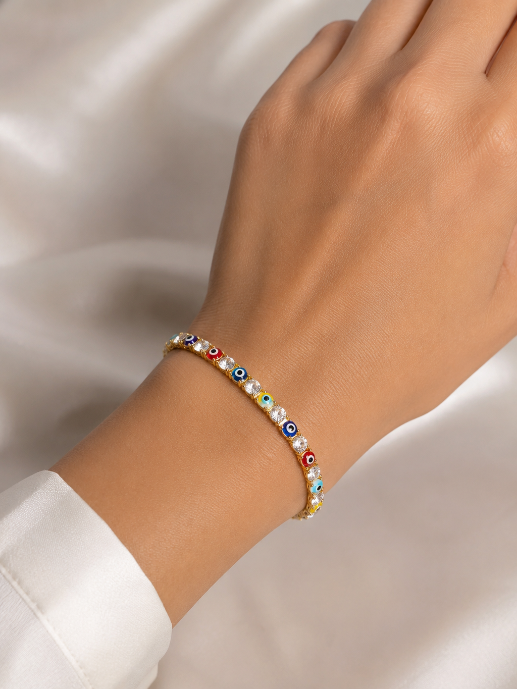 A delicate beaded bracelet with colorful evil eye charms and white pearl-like beads, shown worn on a person's wrist against a soft white satin background.