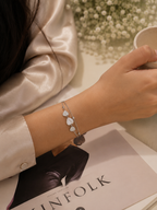 A delicate silver-toned chain bracelet with three white heart-shaped charms and a dark gray stone pendant, worn on a person's wrist. The person is wearing a champagne-colored satin shirt and holding a white ceramic cup, with a magazine and baby's breath flowers in the background.