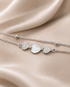 A delicate silver chain necklace with three heart charms, featuring a central white mother-of-pearl heart flanked by two clear crystal-encrusted hearts, displayed on a draped beige satin fabric. Heartstrings Bracelet by Ashaarya featuring a mother-of-pearl heart, American Diamond accents and a lightweight anti-tarnish silver bracelet for women.