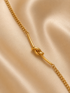 A gold chain necklace featuring a central knotted bar detail, displayed on a draped beige satin fabric background.