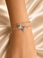 A close-up of a person's wrist wearing two delicate silver bracelets, including a bow-shaped charm bracelet and a thin chain bracelet, set against a draped champagne satin background. Lightweight silver bracelet with American Diamonds by Ashaarya designed for comfortable all-day wear.