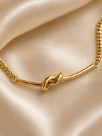 Minimal anti-tarnish gold knot bracelet displayed on a neutral satin background for a luxury jewellery aesthetic.. A gold-toned necklace with a central knotted bar detail and a chain, displayed on draped champagne satin fabric.