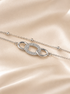 A silver-toned double-strand chain necklace with a central interlocking oval link design, one half of which is paved with small clear stones. The necklace is displayed on a soft, draped champagne-colored satin fabric. The Linked Grace Bracelet by Ashaarya, an anti-tarnish silver bracelet crafted in hypoallergenic 316L stainless steel with sparkling cubic zirconia links for lightweight everyday comfort.