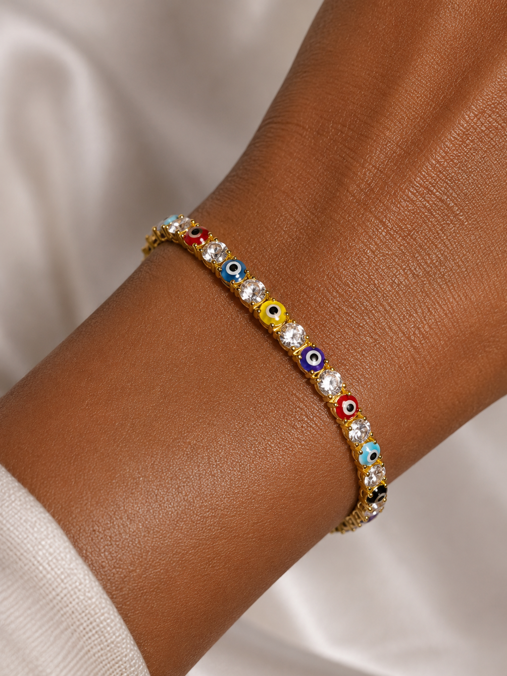 A gold-toned evil eye charm bracelet with clear rhinestones, shown worn on a person's wrist against a soft white fabric background.