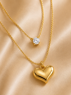 Two gold necklaces displayed on a satin fabric background. The shorter necklace features a single round clear gemstone pendant, while the longer necklace has a polished yellow gold heart-shaped pendant. everyday heart necklace, heart necklace for women, layered necklace for women, gold heart necklace, minimalist gold necklace, anti tarnish necklace, waterproof necklace, 18kt gold plated necklace, stainless steel necklace, dainty layered necklace, everyday jewellery, lightweight necklace, comfort jewellery, 